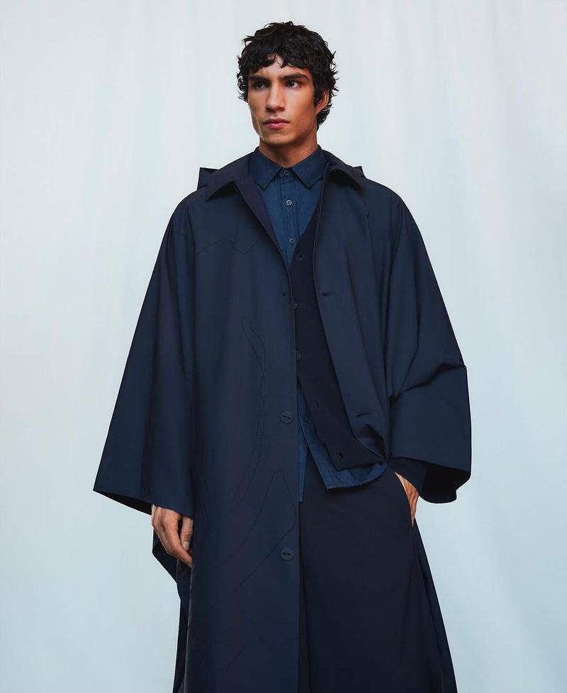 Man in navy overcoat and button-down shirt poses in front of a white background in a fashion or editorial studio shot. 