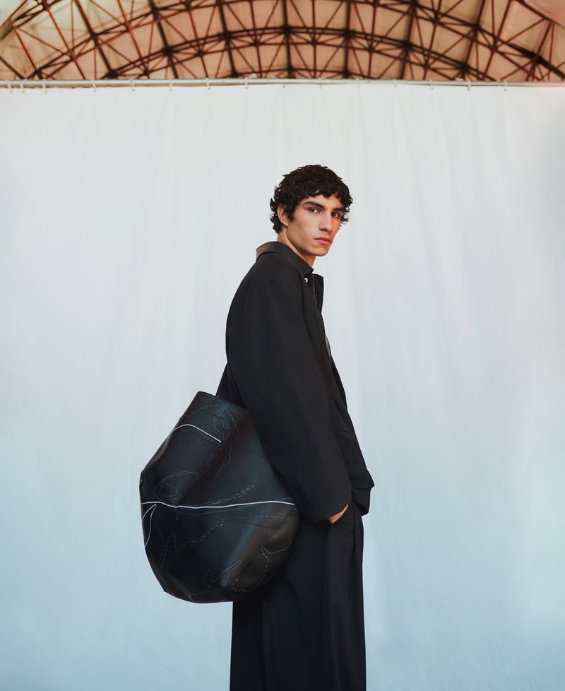 A brunette male fashion model in a black coat poses in front of a large white backdrop with a large black bag hanging on his shoulder.