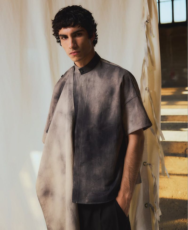 A young adult male poses indoors wearing a grey and black tie-dye short-sleeved shirt with black pants against a neutral backdrop.