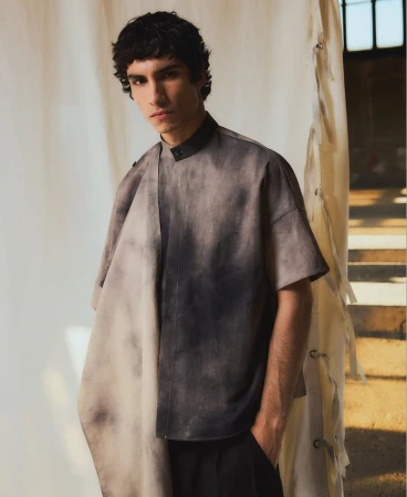 A young adult male poses indoors wearing a grey and black tie-dye short-sleeved shirt with black pants against a neutral backdrop.