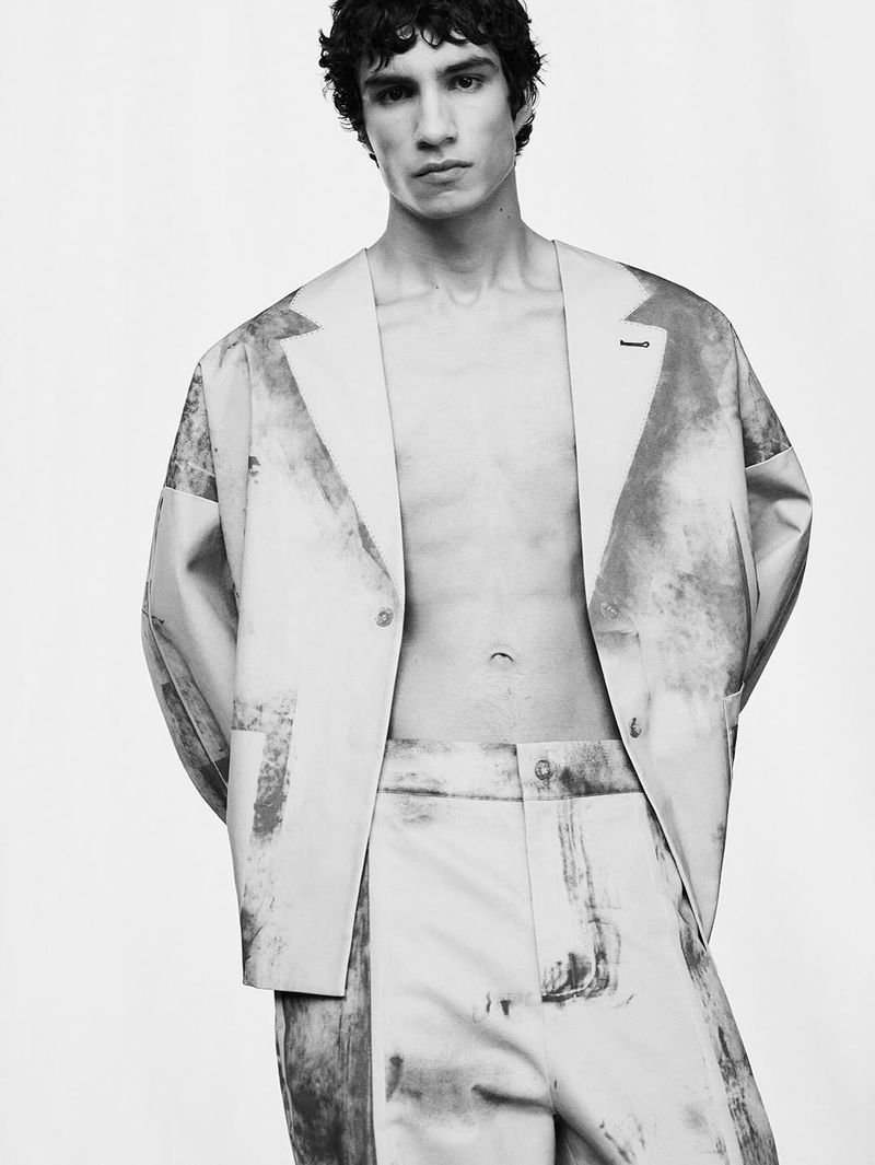 Man poses in a patterned pale-toned jacket and matching pants against a white backdrop in a studio fashion-style shot.