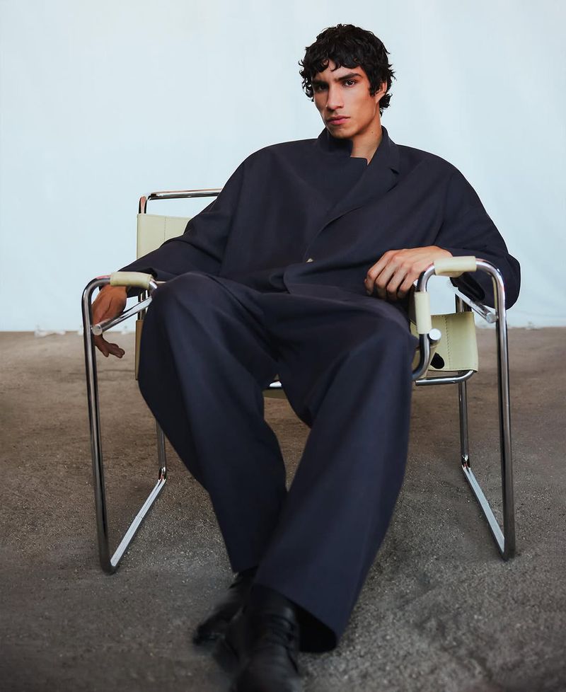 A brunette male model sits in a chrome chair wearing an oversized navy suit in a studio setting with a light blue and gray background.