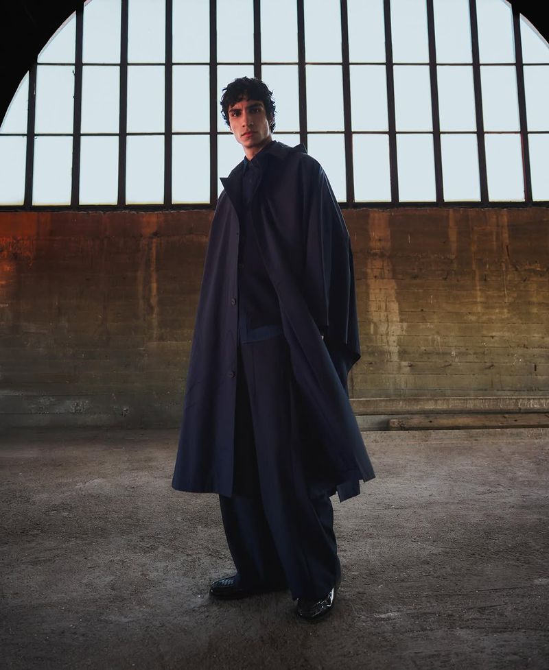 Man poses in a navy trench coat and wide-leg pants indoors. Light from large window reflects on the concrete floor, soft lighting.