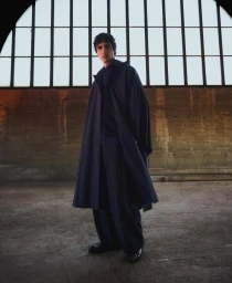Model Duncan Yair wearing an oversized dark navy cape coat and wide-leg trousers in an industrial warehouse setting.