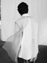 Model seen from behind wearing a white shirt and sheer cape in a studio setting for Adolfo Dominguez F/W 2025