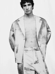 A shirtless male model wearing a patterned suit jacket and trousers against a white seamless background.