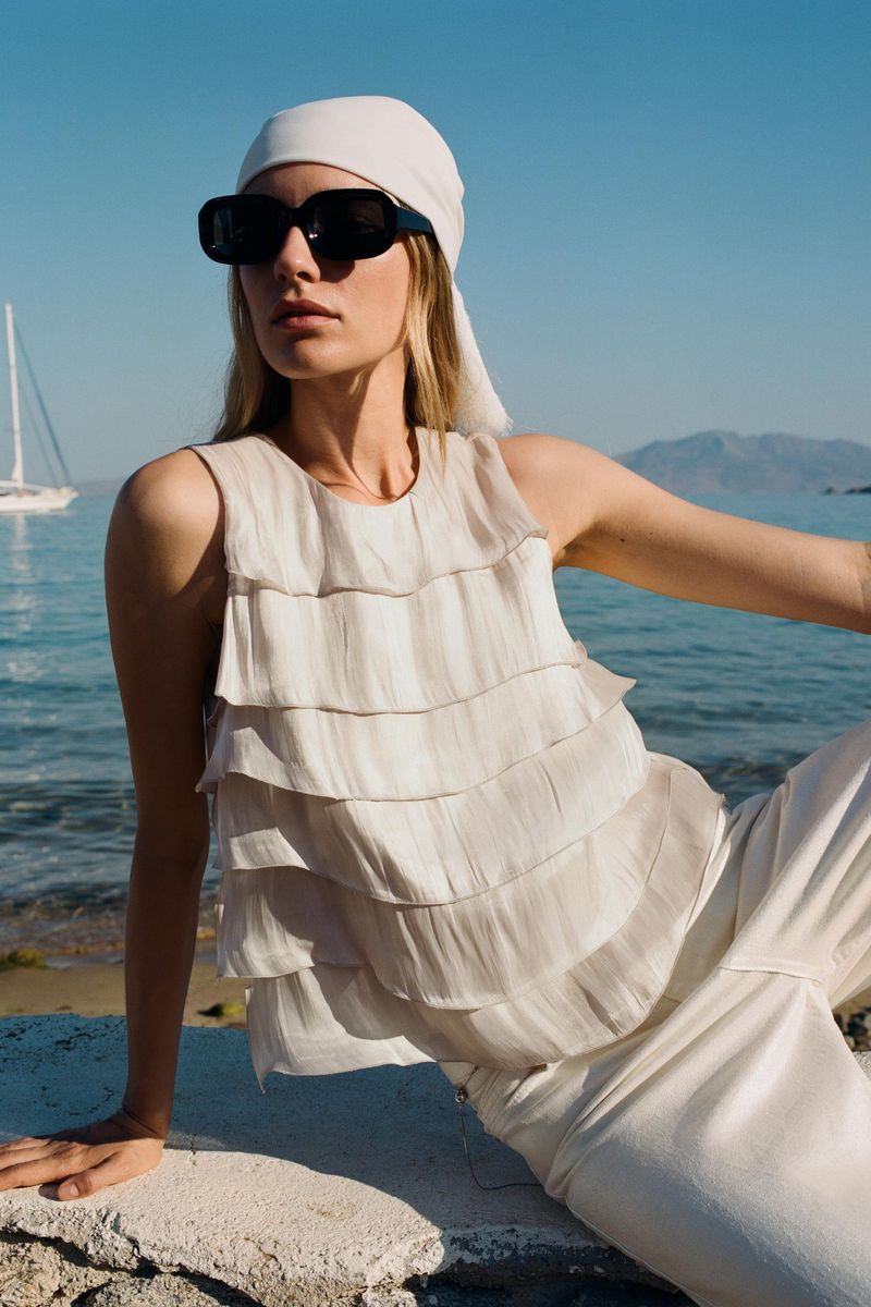 A fair-skinned adult woman wears a cream-colored headscarf, sunglasses, and a layered top while sitting near the sea on a sunny day.