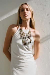 Model wearing a white halter dress and multi-stone necklace posing against a textured white wall in bright sunlight.