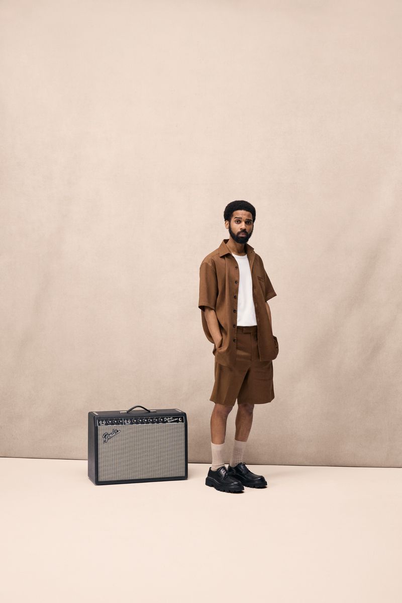 Portrait of a bearded man posing in a matching shirt and shorts set next to a guitar amplifier in a minimalist indoor setting.