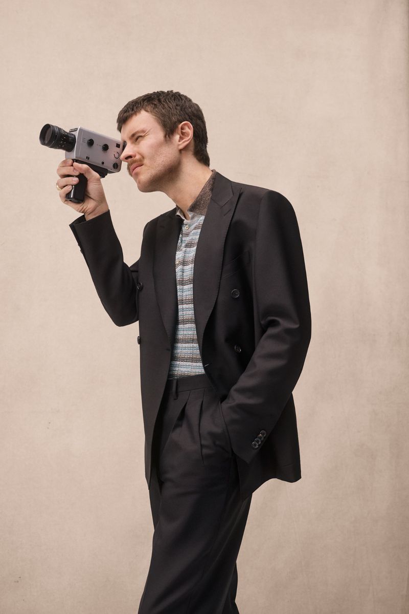 Man in a suit holds a vintage Super 8 film camera, looking through the viewfinder indoors with a neutral background, medium shot.