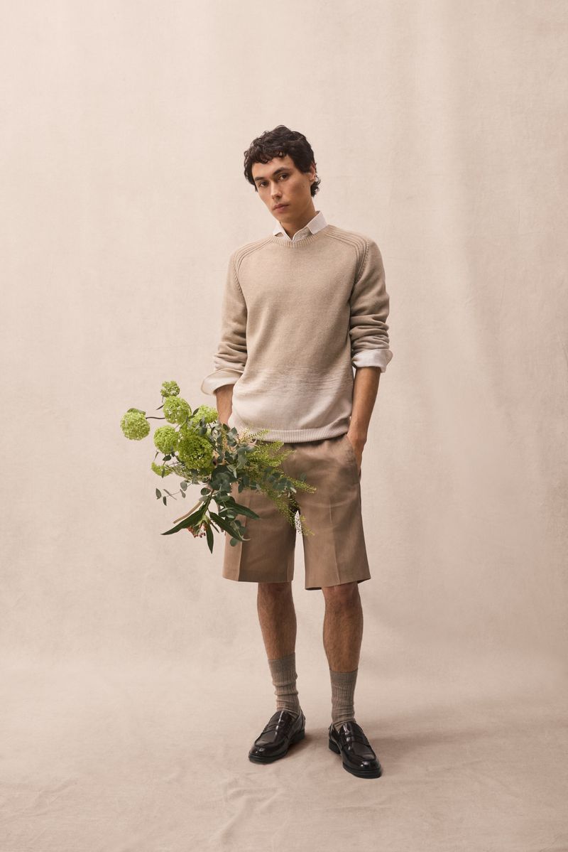 A stylish adult male stands in a light studio setting wearing a sweater, shorts, socks, and loafers while holding green flowers.