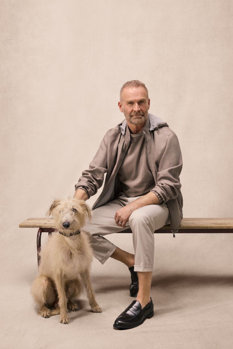 Mature man in a taupe hooded jacket and beige trousers seated on a wooden bench beside a scruffy dog against a neutral studio backdrop.
