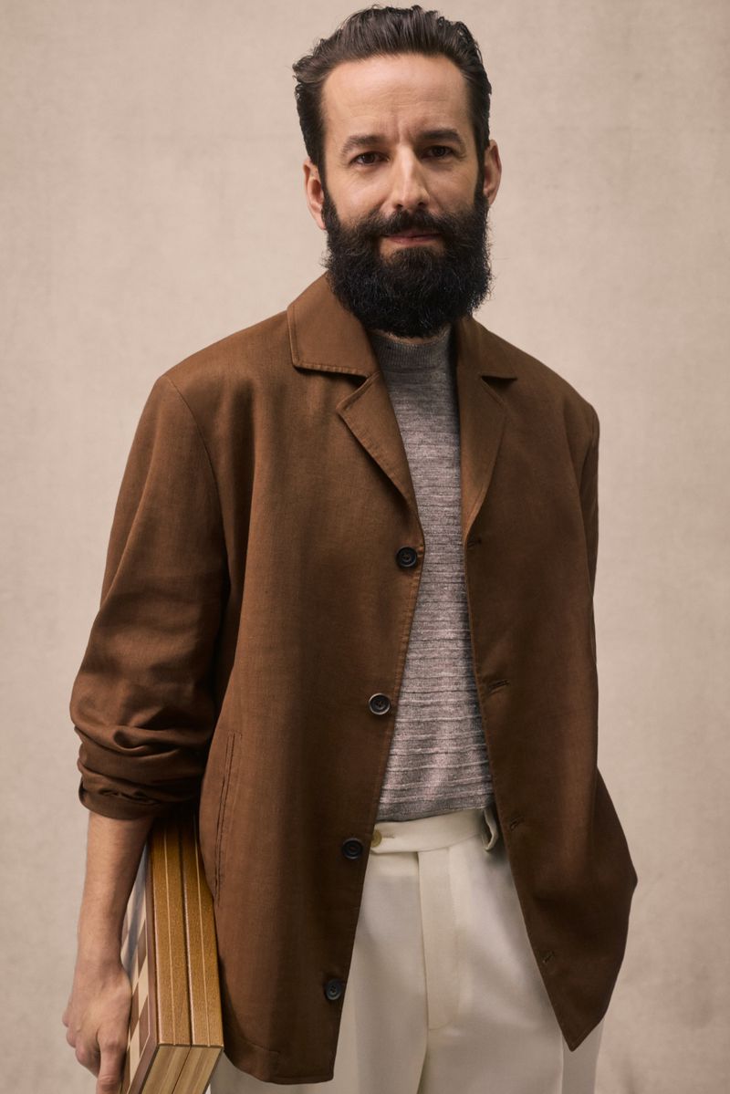 Portrait of bearded man in a brown jacket holding a chessboard against a neutral backdrop. Studio shot, looking at camera.