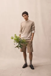 Male model wearing a beige knit sweater and tailored shorts holding a green floral bouquet against a neutral fabric studio backdrop.