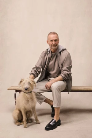 Mature man in a taupe hooded jacket and beige trousers seated on a wooden bench beside a scruffy dog against a neutral studio backdrop.