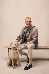 Mature man in a taupe hooded jacket and beige trousers seated on a wooden bench beside a scruffy dog against a neutral studio backdrop.