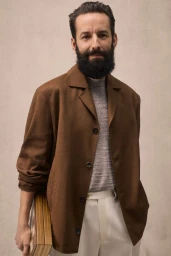 Man with full dark beard wearing a brown jacket over a grey knit sweater and white trousers against a neutral background