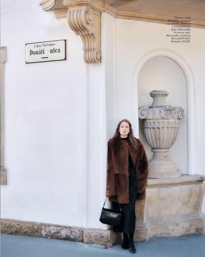 Model wearing a brown fur coat and black skirt posing outdoors against a white wall with an arched stone urn niche.
