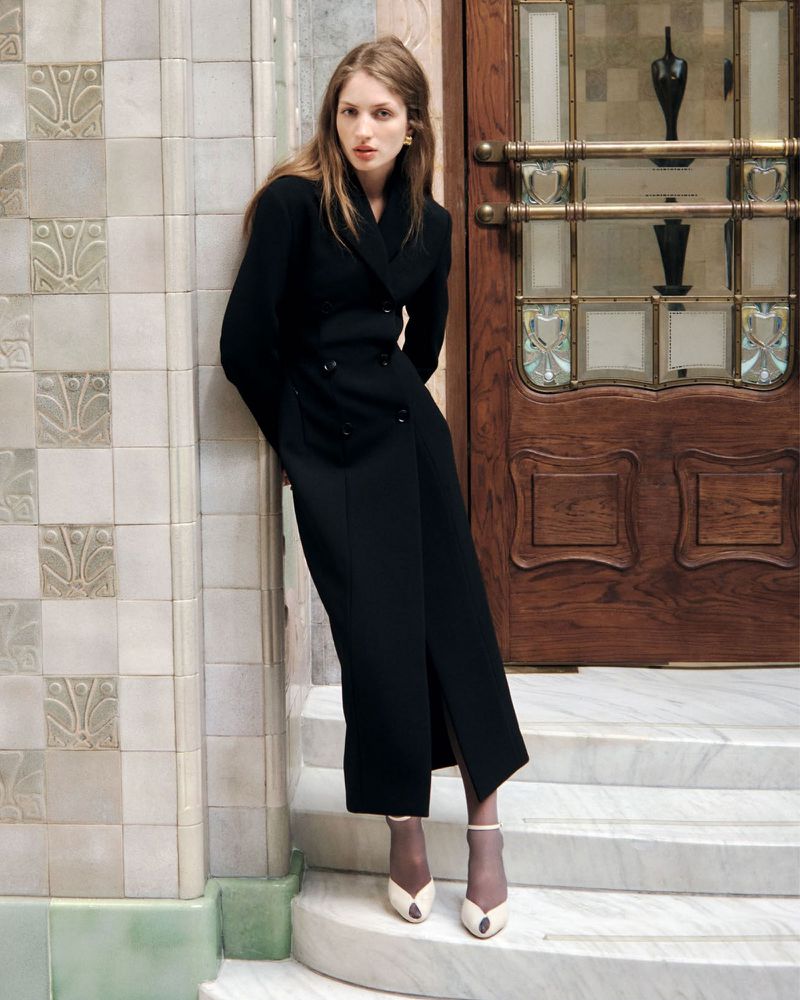 Model in a long black double-breasted coat leaning against a patterned tile wall next to a wooden doorway