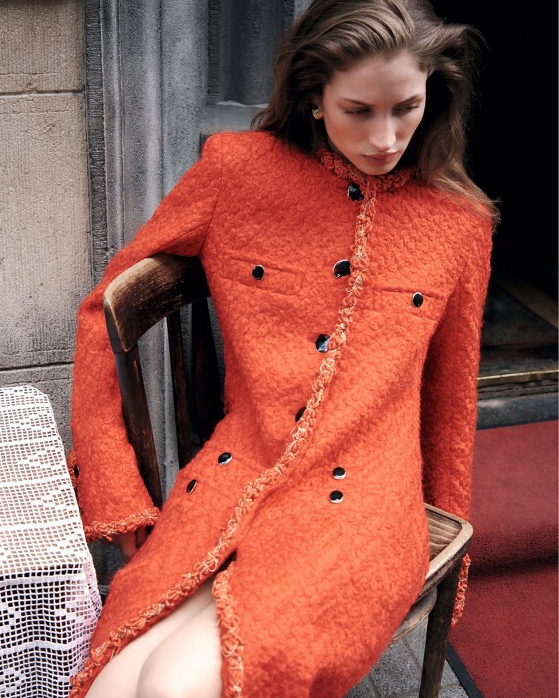 Model wearing a textured orange jacket seated on a wooden chair outdoors near a doorway