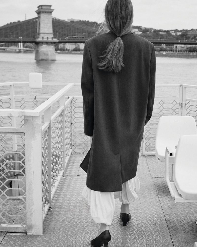Model seen from behind wearing a long dark coat walking on a boat deck with a bridge in the background.