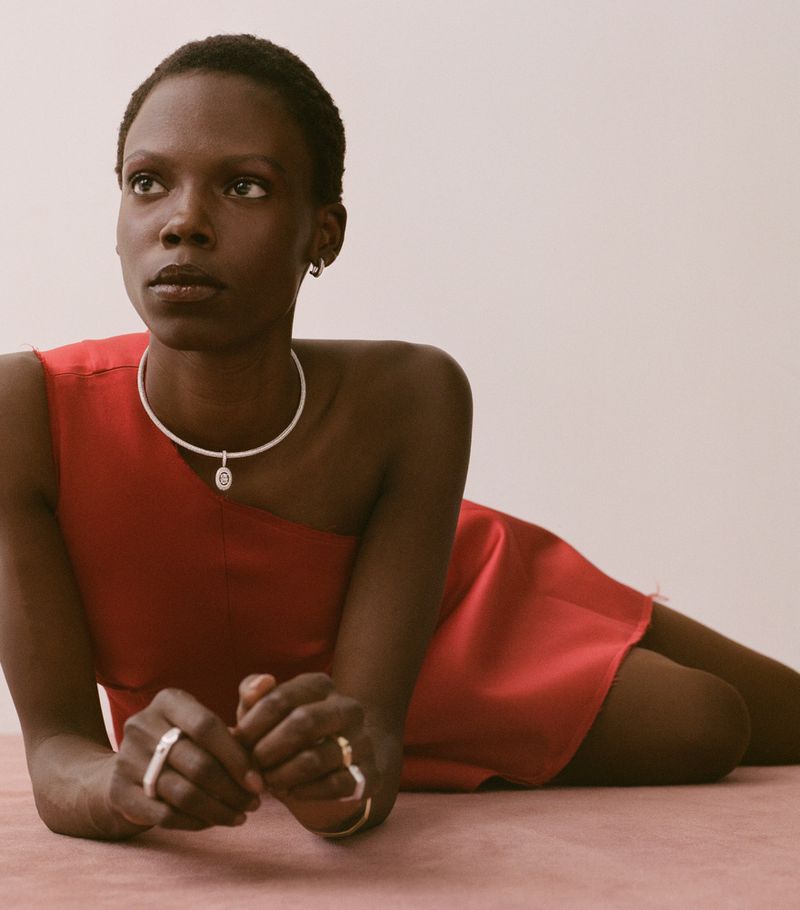 Model Agel Akol in a red dress wearing silver jewelry lying against a neutral background for Heroine Magazine