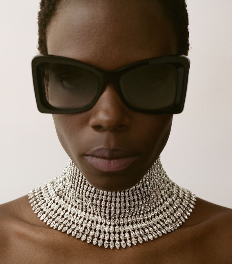 Close-up portrait of model wearing oversized black sunglasses and a wide crystal choker necklace on a white background
