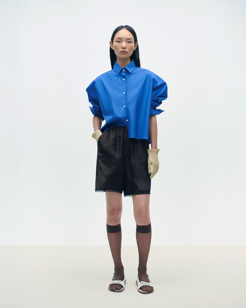 Fashion model in a blue button-down shirt, black shorts, sheer socks, and sandals poses in a brightly lit photo studio.