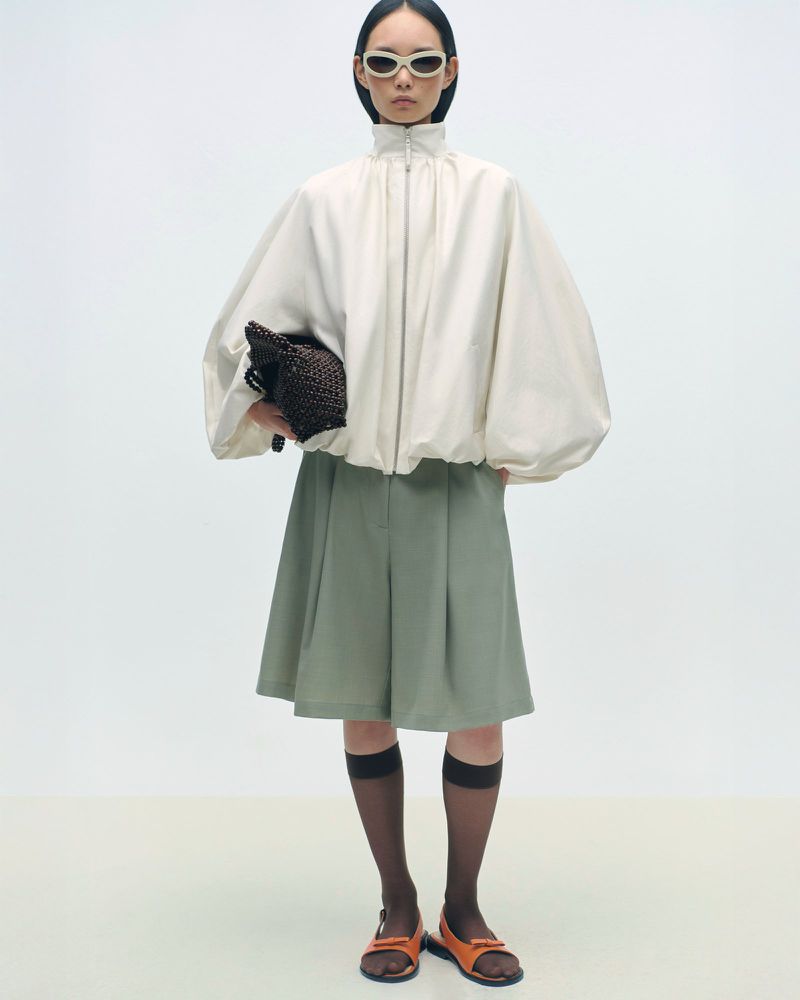 Fashion shot of a woman in sunglasses wearing a large beige jacket, green culottes, socks, and sandals holding a black woven purse.