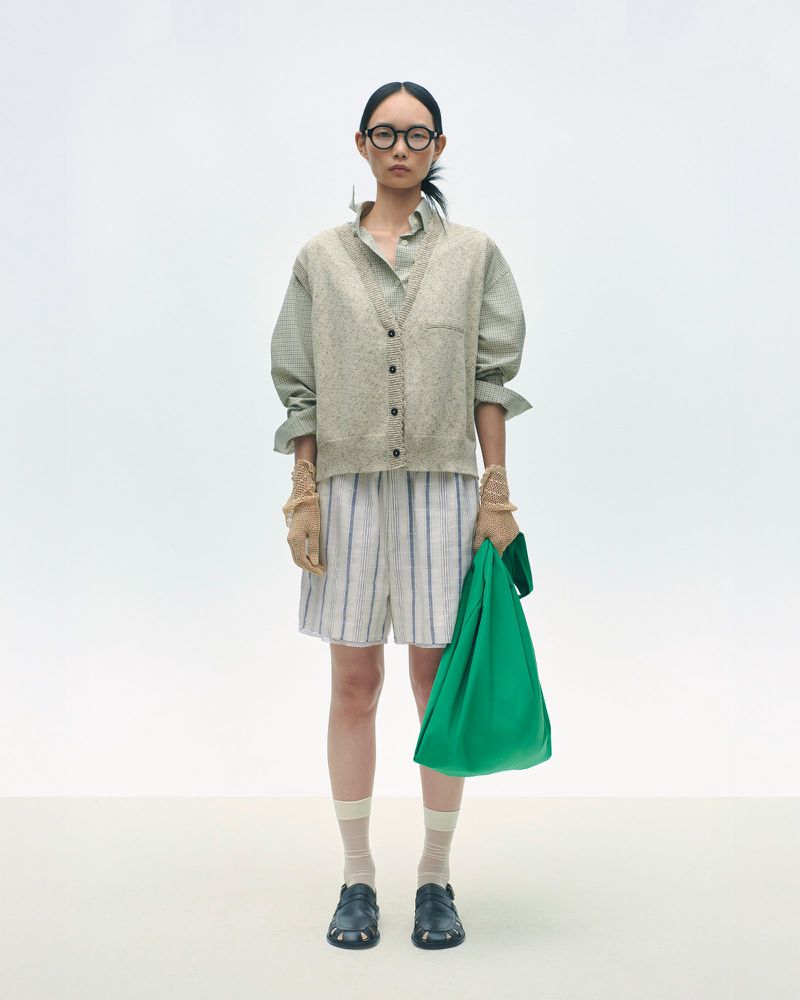 Adult woman in glasses posing in neutral palette clothing and holding a green tote bag in a brightly lit white photo studio.