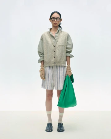 Adult woman in glasses posing in neutral palette clothing and holding a green tote bag in a brightly lit white photo studio.