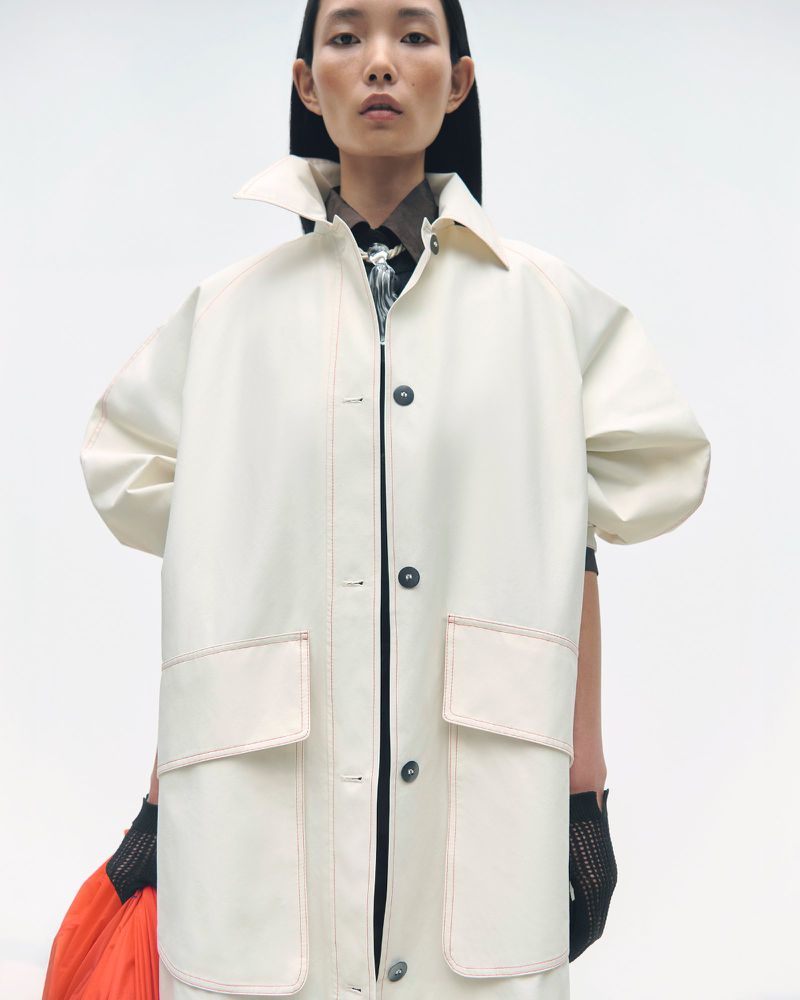 An Asian woman models an oversized white trench coat with orange details against a clean, bright background in a studio fashion shot.