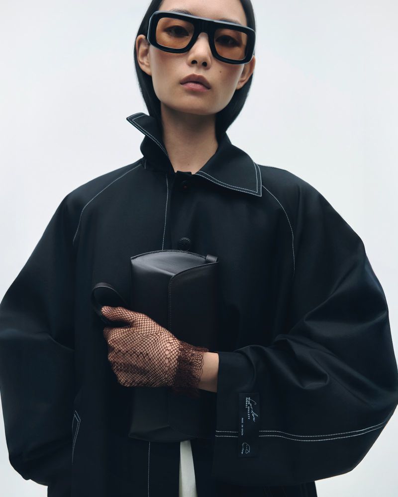 Asian woman posing in a black coat and black glasses while holding a structured handbag against a neutral backdrop in a studio setting.