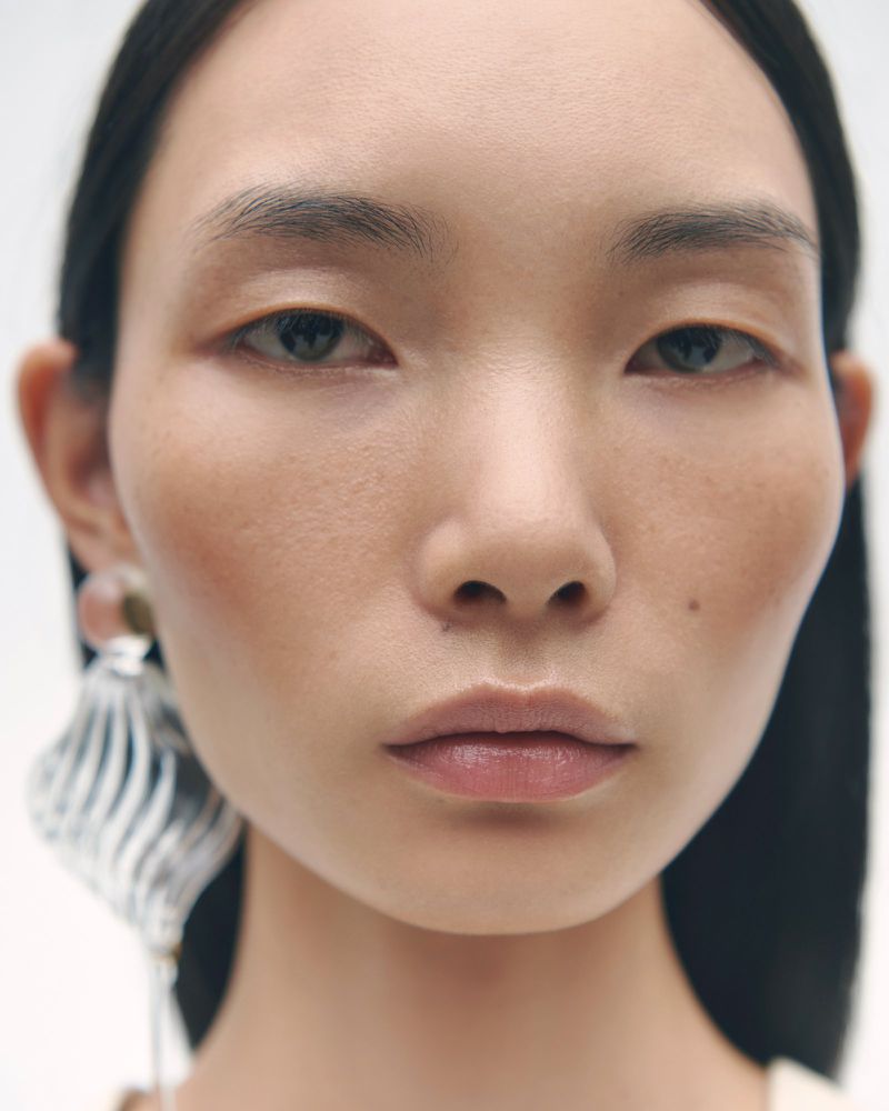Close-up portrait of model Ling Chen wearing a sculptural silver statement earring against a white background.