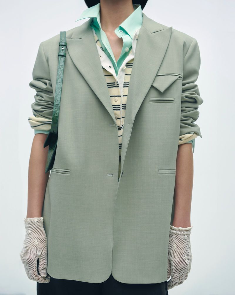 Model wearing a green blazer and gloves stands in a studio setting. The composition is a medium shot focused on the upper body.