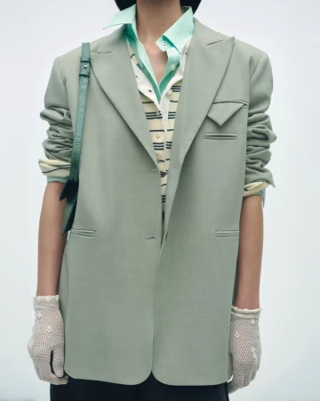 Model wearing a green blazer and gloves stands in a studio setting. The composition is a medium shot focused on the upper body.