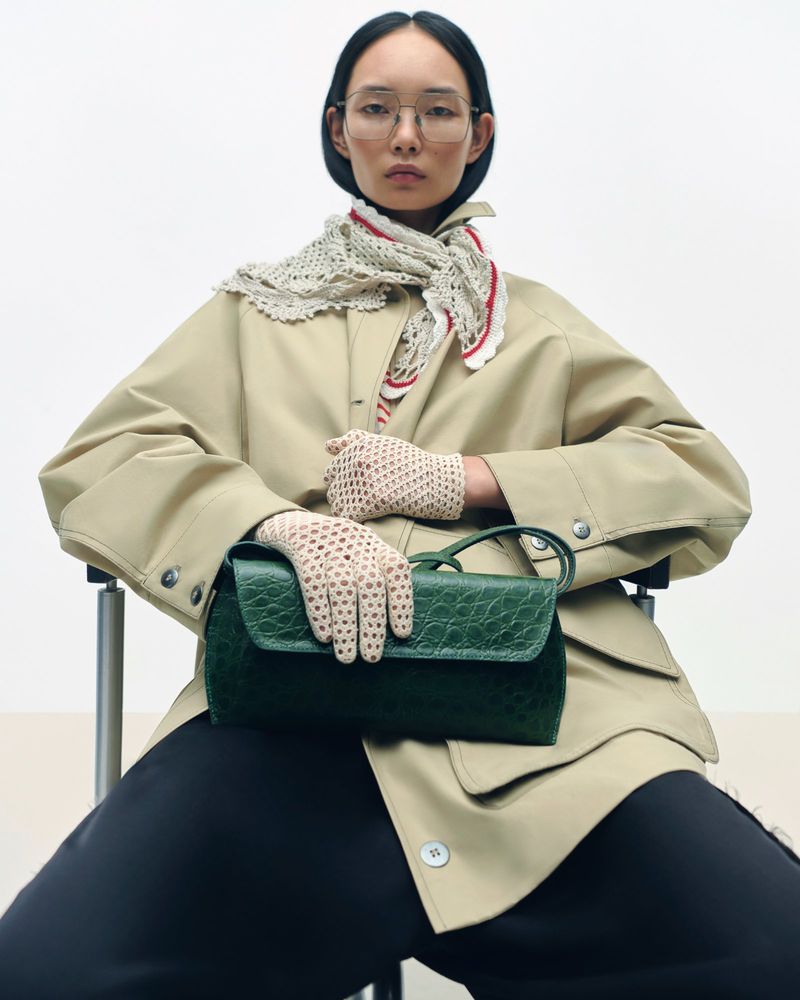 A fashion shot of a woman wearing a trench coat, glasses, scarf, and gloves holding a green leather handbag against a bright background.