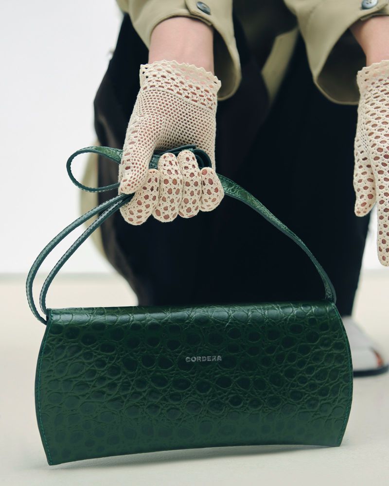 Close-up of a gloved hand holding a dark green handbag with a crocodile texture against a white backdrop in a studio setting.