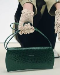 Cropped view of hands in off-white crochet gloves holding a dark green crocodile-embossed leather Cordera shoulder bag.