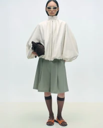 Model wearing a white high-neck jacket and green shorts holding a dark handbag against a seamless white studio background.