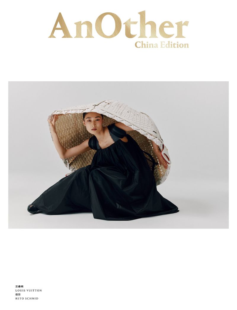 Model seated on a white floor holding a large woven structured hat over her head while wearing a black dress.