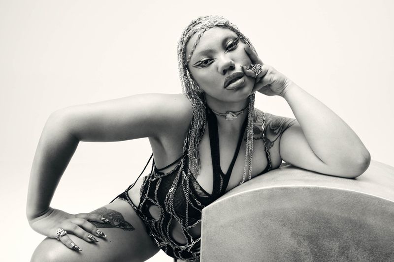 Monochromatic portrait of a woman with elaborate chain headdress, heavy makeup, and bodysuit, leaning on a textured block in a studio.