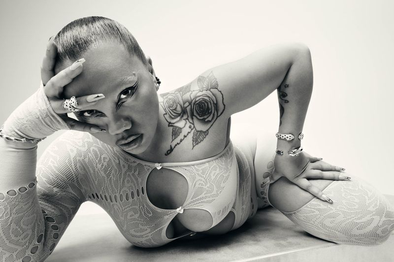 Close-up black and white shot of a woman in a lace bodysuit, face framed by her hands. She is lying down, posing for the camera.
