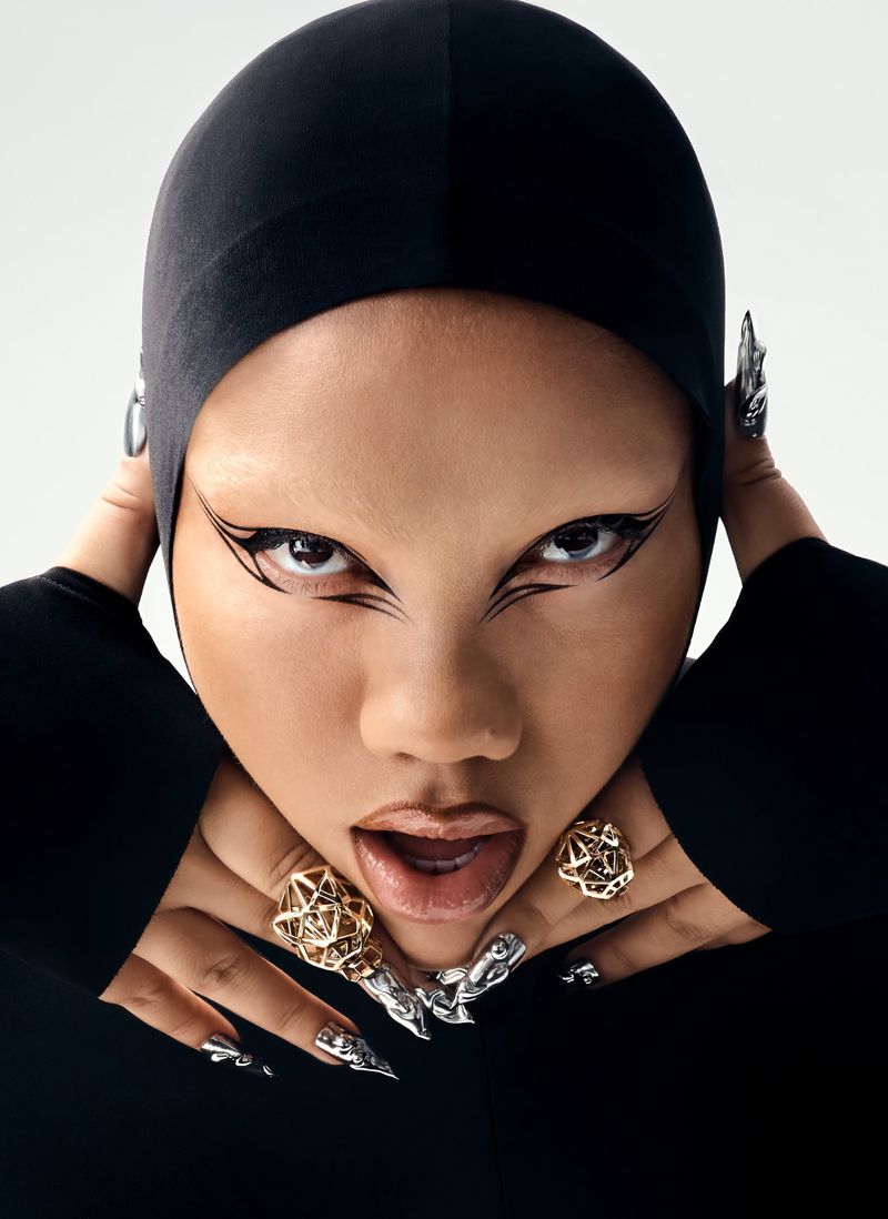 Close-up portrait of Parris Goebel wearing a black cap, dramatic graphic eyeliner, gold rings, and metallic sculptural nail art.