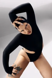 Parris Goebel wearing a black bodysuit and hood in a dynamic pose against a white studio background.