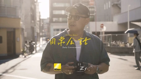 Man wearing a cap and glasses holding a camera on a city street for Nikon