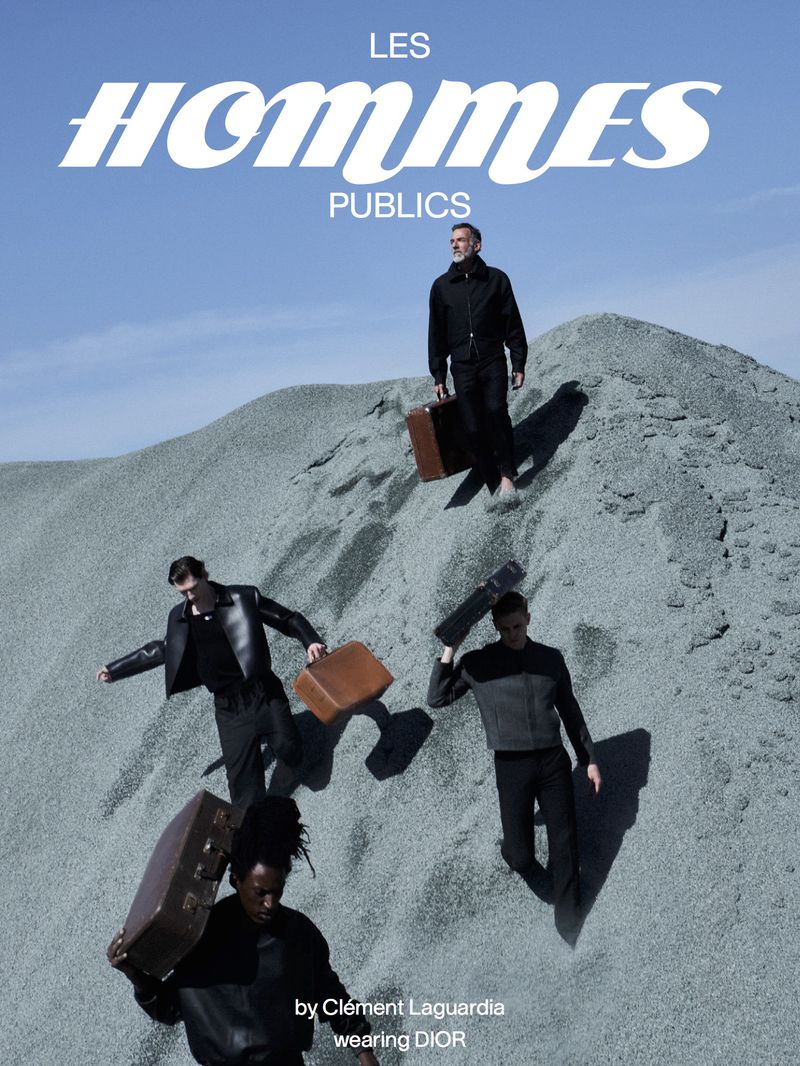 Four men in fashionable dark attire are on a sandy mound carrying briefcases against a light blue sky for a fashion editorial.