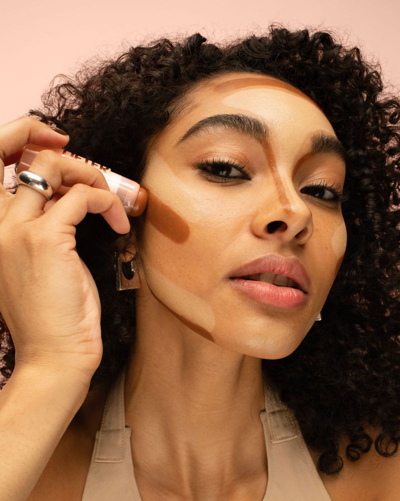 Woman applying face makeup with a stick. Close-up shot of an adult female using a light brown stick on her face, with curly hair.
