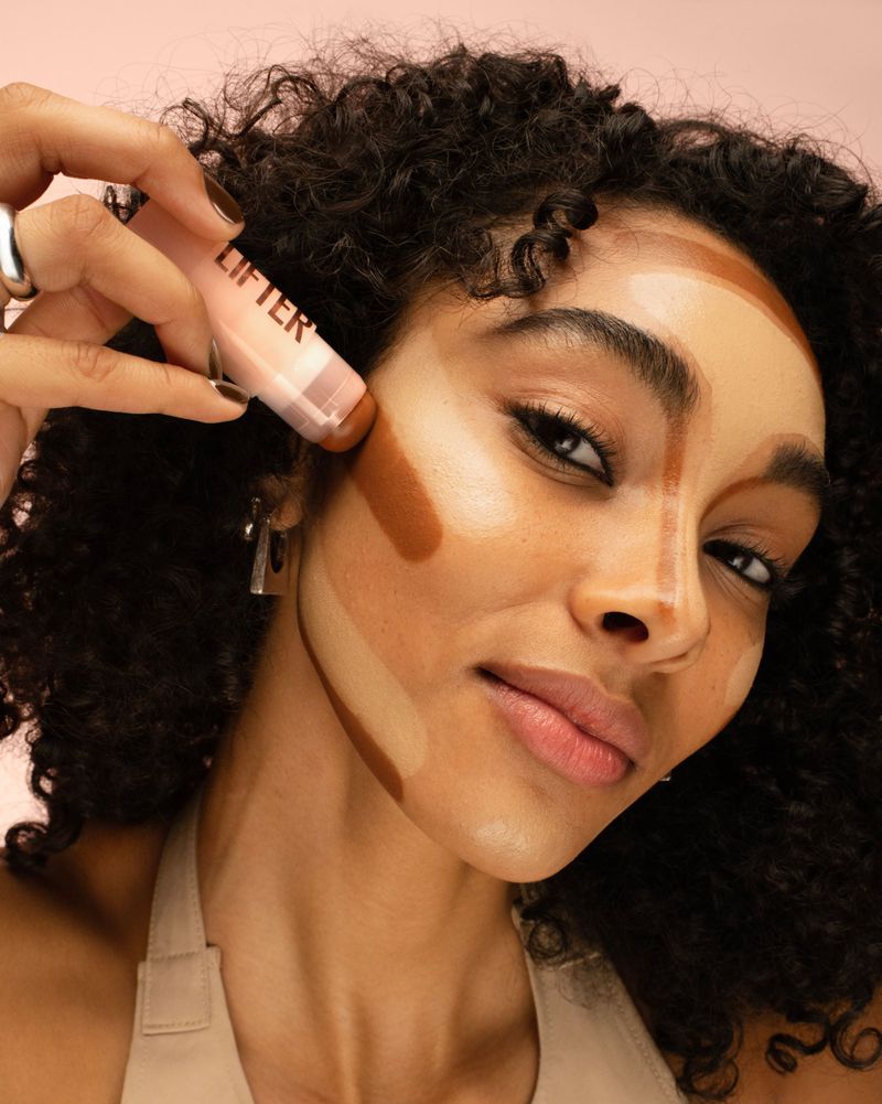 Close-up of a woman with medium skin tone applying contouring makeup to her face using a makeup applicator stick, in a beauty shot.