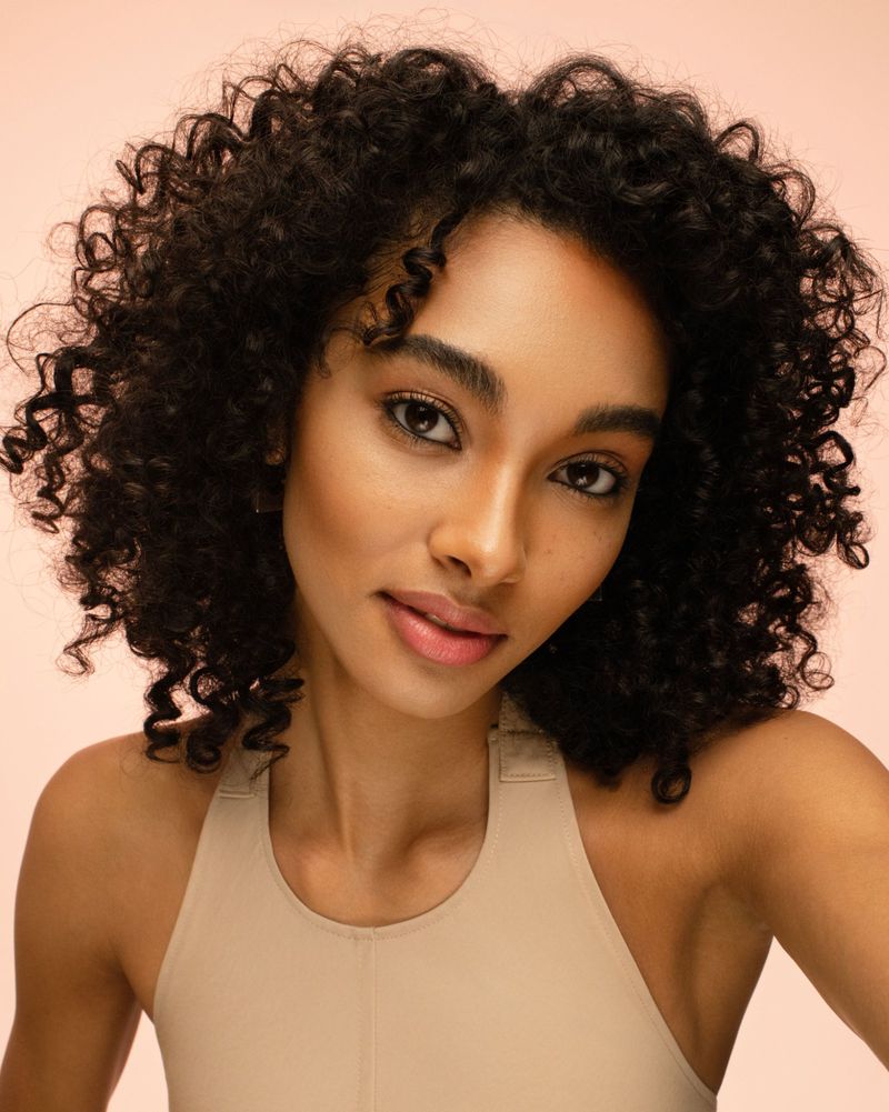 Close-up of a young adult woman with dark curly hair, wearing a beige top, smiling slightly against a peach-colored background.